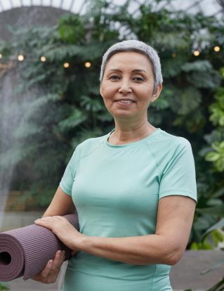 Elderly woman holding yoga mat and smiling in a lush greenhouse setting.